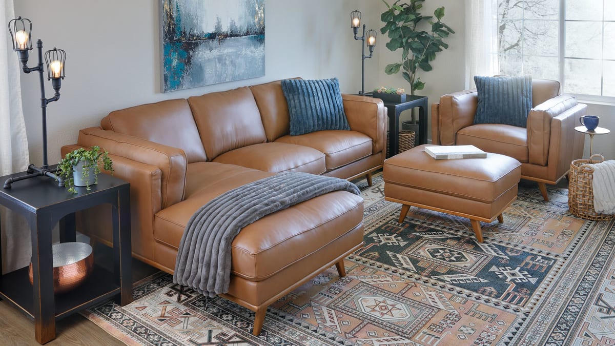 Camel colored brown leather sectional accompanied by matching chair and ottoman in blue and brown toned living room window scene