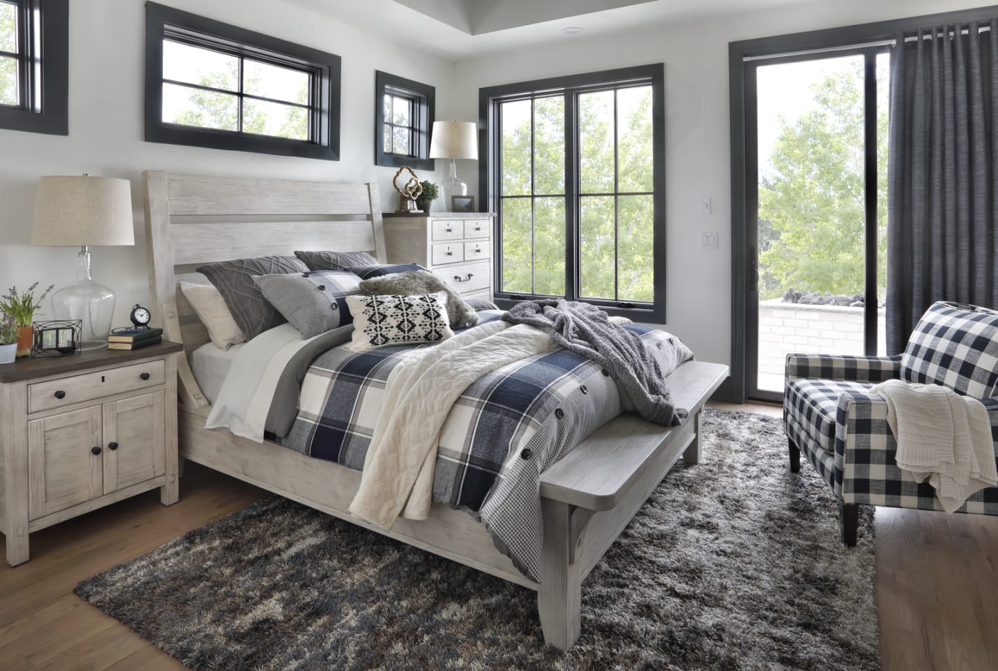 Cream colored farmhouse bedroom set with slatted headboard accompanied by matching nightstand and chest in bedroom window scene