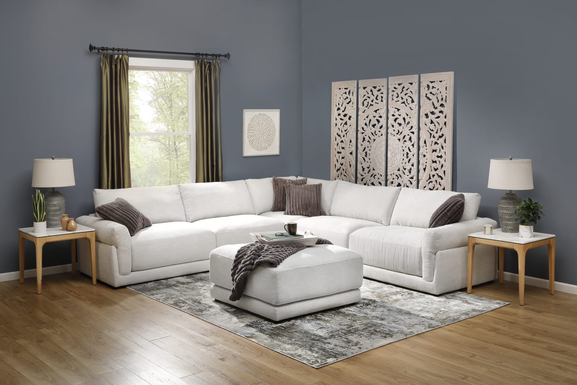 A living room window scene featuring the Lennox Sectional and Ottoman accompanied by organic modern home decor like end tables, lamps, and woven wall art