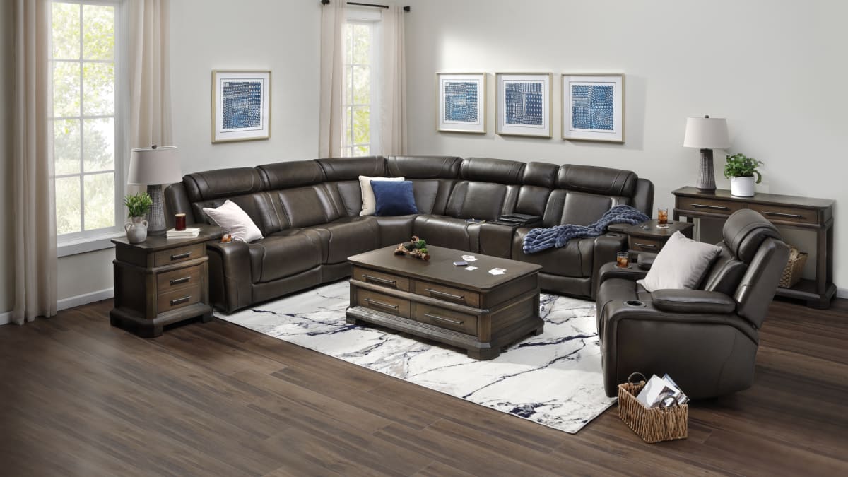 A dark brown leather sectional in a living room with a hardwood floor and white rug. Also shows a reclining chair and coffee table